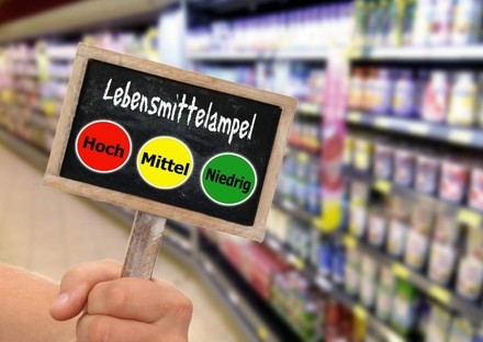 Eine Hand hält in einem Supermarkt ein Schild hoch auf dem die Lebensmittelampel zu sehen ist. Rot für hoch, gelb für mittel und grün für niedrig
