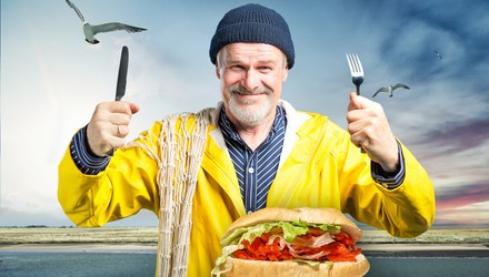 Ein Fischer hält Messer und Gabel hoch um ein vor ihm liegendes Fischbrötchen zu essen