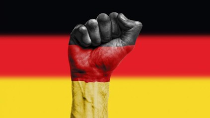 Eine Hand zur Faust geballt in Deutschlandfarben angemalt, im Hintergrund die Deutschlandflagge