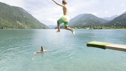 Ein Mann springt von einem Sprungbrett in einen See in dem bereits ein anderer Mann schwimmt