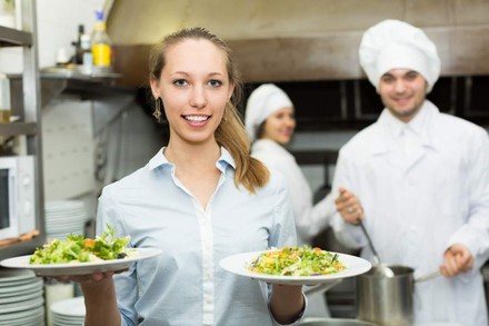 Eine Serviekraft mit zwei Tellern Salat, im Hintergrund zwei Köche bei der Arbeit