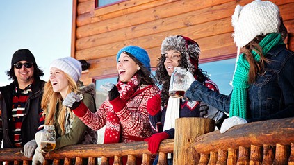 Fünf Personen stehen auf einer Terrasse einer Skihütte in Winterbekleidung und lachen