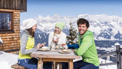 Eine Gruppe Wintersportler sitzt am Tisch vor einer Hütte und isst gemeinsam