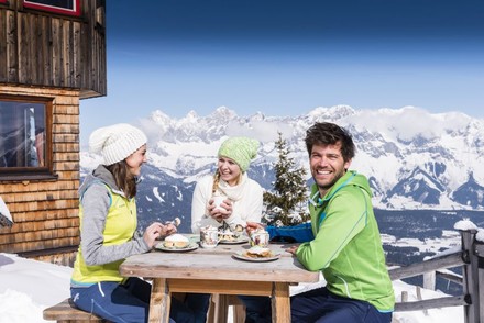 Eine Gruppe Wintersportler sitzt am Tisch vor einer Hütte und isst gemeinsam