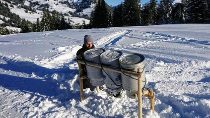 Ein Mann kniet im Schnee vor drei großen Milchkannen, die dort aufgestellt wurden