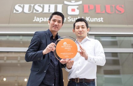 Zwei Asiaten stehen vor dem Sushi Plus Restaurant und halten den Preis für Best Restaurants Awards 2017 hoch