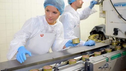 Eine Mitarbeiterin überwacht das Fließband einer Dosenwurstfabrik, ihr Kollege arbeitet im Hintergrund an einer Maschine