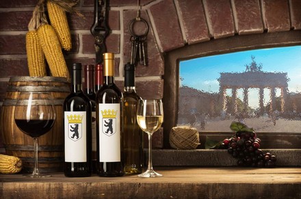 Mehrere Weinflaschen und Weingläser stehen auf einer Holzanrichte, im Hintergrund ist das Brandenburger Tor durch ein Fenster zu erkennen