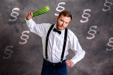 Eine junger Mann schwingt einen Zucchini wie einen Knüppel, im Hintergrund sind Paragraphenzeichen zu sehen