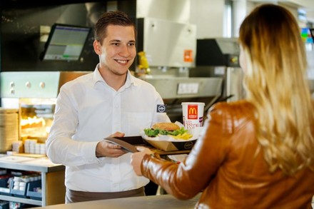 Das Essen kann man natürlich weiterhin selbst am Counter abholen, künftig wird es aber auch an den Tisch geliefert. (© McDonald’s Österreich)
Ein freundlicher McDonald’s Mitarbeiter händigt einer Kundin ein Tablett mit Burger und Getränk aus