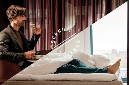 Ein Mann sitzt an einem Tisch beim Essen, seine Beine jedoch befinden sich bereits im Bett. Davor steht Scandic Friends