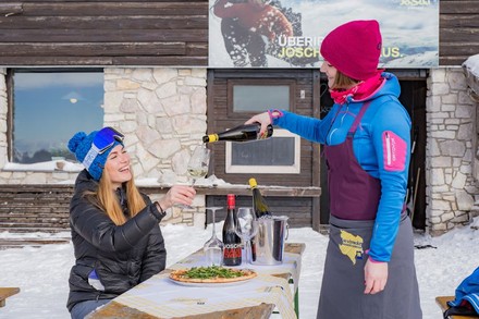 Eine junge Servicekraft schenkt einer Wintersportlerin auf einer Hütte im Schnee Wein nach