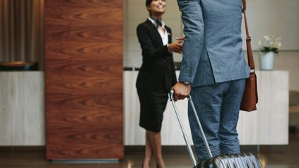 Ein Mann im Anzug und mit Koffer wird von einer Hotelmitarbeiterin in einer Hotellobby per Handschlag begrüßt