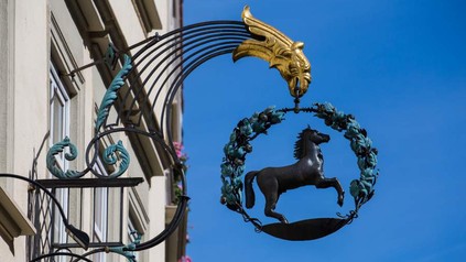 Ein Eisengestell das ein Pferd in einem Lorbeerkranz, gehalten von einem goldenen Vogelkopf zeigt hängt an einer Hauswand