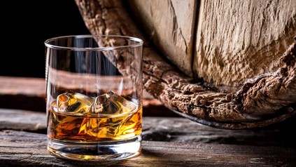 Ein Glas mit Whiskey und Eiswürfeln vor einem Holzfass