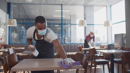 Damit die Gäste in Restaurants trotz Hygienekonzepte nicht ausbleiben, denkt NRW jetzt über Luftfilter für die Gastronomie nach. (Foto: ©Anatoliy/stock.adobe.com)
Afrikanischer Restaurantmitarbeiter mit Maske und Handschuhen reinigt einen Tisch
