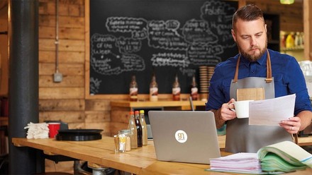 Junger Gastronom mit Ausdrucken und Kaffee in dr Hand vor seinem Laptop