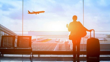 Eine Person steht mit Kaffee und Koffer in einer Wartehalle am Flughafen und sieht einem Flugzeug beim Abheben zu