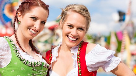 In Tracht aufs Frühlingsfest nach München! (Foto: © Kzenon / stock.adobe.com) Zwei junge Frauen im Dirndl stehen auf der Theresienwiese. Im Hintergrund sind Fahrgeschäfte.