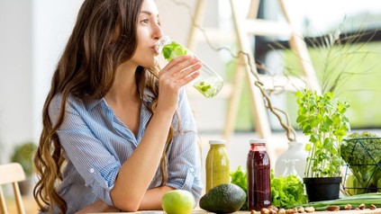 Eine frau sitzt an einem Tisch mit Smoothies, einer Avocado und einem Apfel und trinkt aus einer Flasche mit Wasser und Kräutern