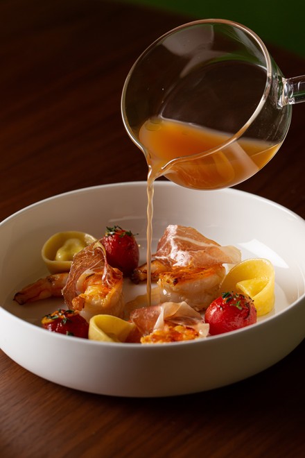 Tiefer weißer Teller der gekochte mit Schinken bedeckte Garnelen, hausgemachte Nudeln und ganze Tomaten enthält. Klare, bernsteinfarbene Soße wird aus einer durchsichtigen Kanne in den Teller gegossen.