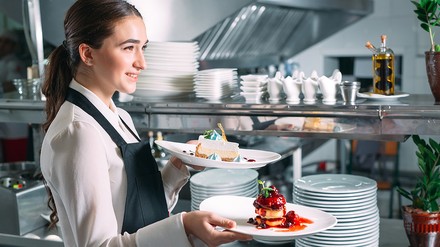 Eine weibliche Bedienung steht am Küchenpass und trägt Dessert auf weißen Tellern zum Gast.