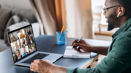 Ein Mann sitzt vor seinem aufgeklappten Laptop. Er nimmt an einem Zoom-Meeting teil. Auf dem Display seines Rechners sind  andere Teilnehmer des Meetings zu sehen.