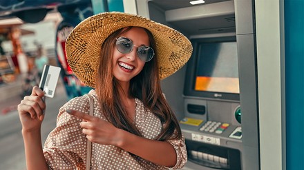 Zahlen bitte! – Mit Cash oder Karte? (Foto: © Valerii Apetroaiei/stock.adobe.com) Eine junge Frau mit Sommerhut steht vor einem Geldautomat und hebt ihre Kreditkarte in die Kamera.