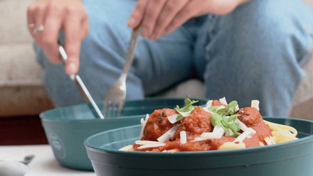 Hände eines sitzenden Mannes mit Besteck, der aus einer Mehrwegverpackung Spaghetti Bolognese isst