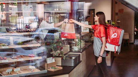 Junge Frau holt in Namen von Delivery Hero Speisen in einer Tüte in einem Restaurant ab.  (Foto: © Delivery Hero)