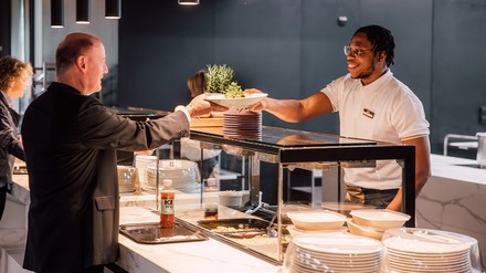 Ein junger Gastromitarbeiter reicht einen etwas älteren Herren einen Teller mit Salat.