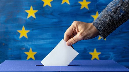 Eine Hand steckt einen Wahlzettel in einen Karton, im Hintergrund die Fahne der EU