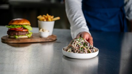 Eine Hand schiebt einen Salat mit Mayo in den Vordergrund, im Hintergrund steht ein Burger mit Pommes