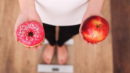 Eine Frau steht auf einer Waage und hält in einer Hand einen Apfel, in der anderen einen Donut