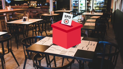 Leeres, abgedunkeltes Restaurant mit dunklen Holzmöbeln und einem Gasttisch auf dem eine rote Wahlurne steht. Es wird ein Stimmzettel eingeworfen. Auf dem Stimmzettel ist der Schriftzug der SPD zu lesen, darunter das ausgefüllte Wahlkreuz.