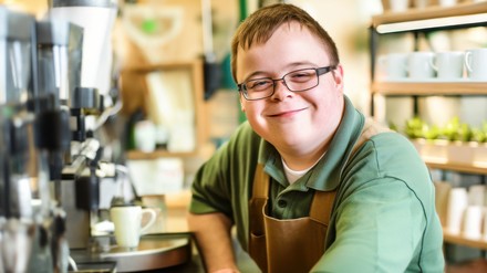 Junge Mann mit Chromosomenanomalie steht vor eine Barista-Kaffeemaschine in einem Cafe