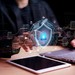 Mann sitzt vor Laptop und Tablet. Zeigt mit Finger auf Sicherheits-Symbol.