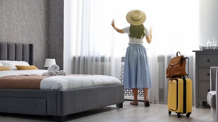 Urlauberin steht im Hotelzimmer und schaut aus dem Fenster, neben ihr steht ihr Koffer und ihr Rucksack