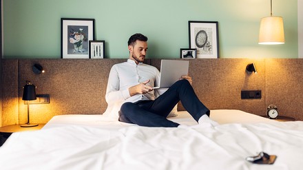 Mann sitzt auf einem Hotel-Bett und arbeitet mit seinem Laptop