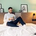 Mann sitzt auf einem Hotel-Bett und arbeitet mit seinem Laptop
