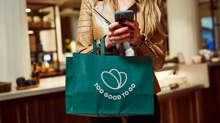 Frau mit Smartphone und Too Good To Go Tüte