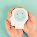Zwei Hände halten einen symbolischen Kopf, im Gehirn ein lachender Smiley
