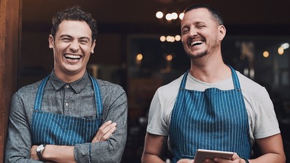 Zwei Mitarbeiter aus der Gastronomie stehen lachend im Eingangsbereich eines Betriebs und tragen Arbeitsschürzen