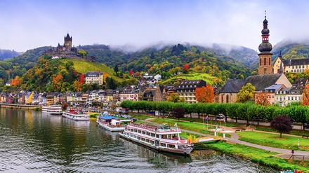 Flusskreuzfahrtschiffe auf einem Fluss in Rheinland-Pfalz vor einer Stadt mit Kirche und Burg.