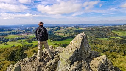 Wanderer steht auf einem Felsen am Pferdskopf an der Wasserkuppe in der Rhön und blickt über eine weite Landschaft aus Feldern und Wäldern.