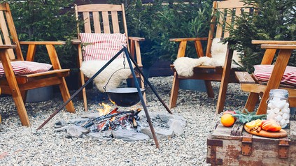 Gemütlicher Außenbereich mit Holzstühlen, Decken und Feuerstelle; über dem Feuer hängt ein Topf, daneben liegen winterliche Snacks und Getränke.