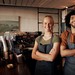 Zwei Mitarbeiter stehen lächelnd hinter der Theke eines Cafés und tragen Schürzen; im Hintergrund sind Kaffeemaschine und Gläserregal zu sehen.