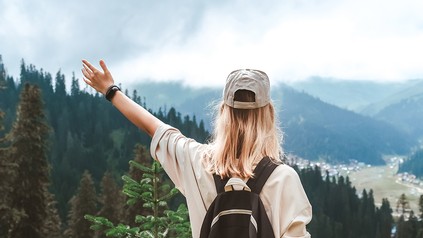 Junge Reisende mit Rucksack blickt von einem Bergpfad auf ein Tal mit Wald und Bergen