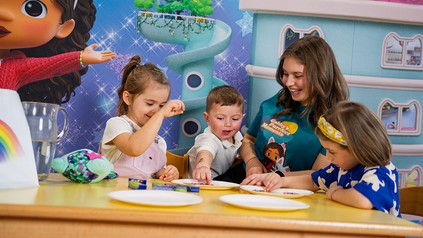 Kinder spielen in einem Kinderhotel gemeinsam mit einer Betreuungsperson am Tisch mit Figuren aus „Gabby’s Dollhouse“