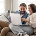 Paar sitzt entspannt auf einem Sofa und nutzt gemeinsam einen Laptop, um online eine Reise zu planen.